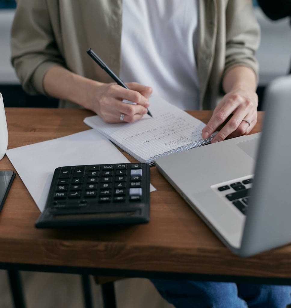 Person at a laptop writing on a notepad alongside a black calculator